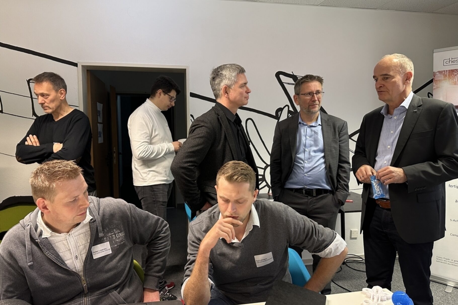 3 Männer unterhalten sich, Im Vordergrund sitzen 2 Männer an einem Tisch