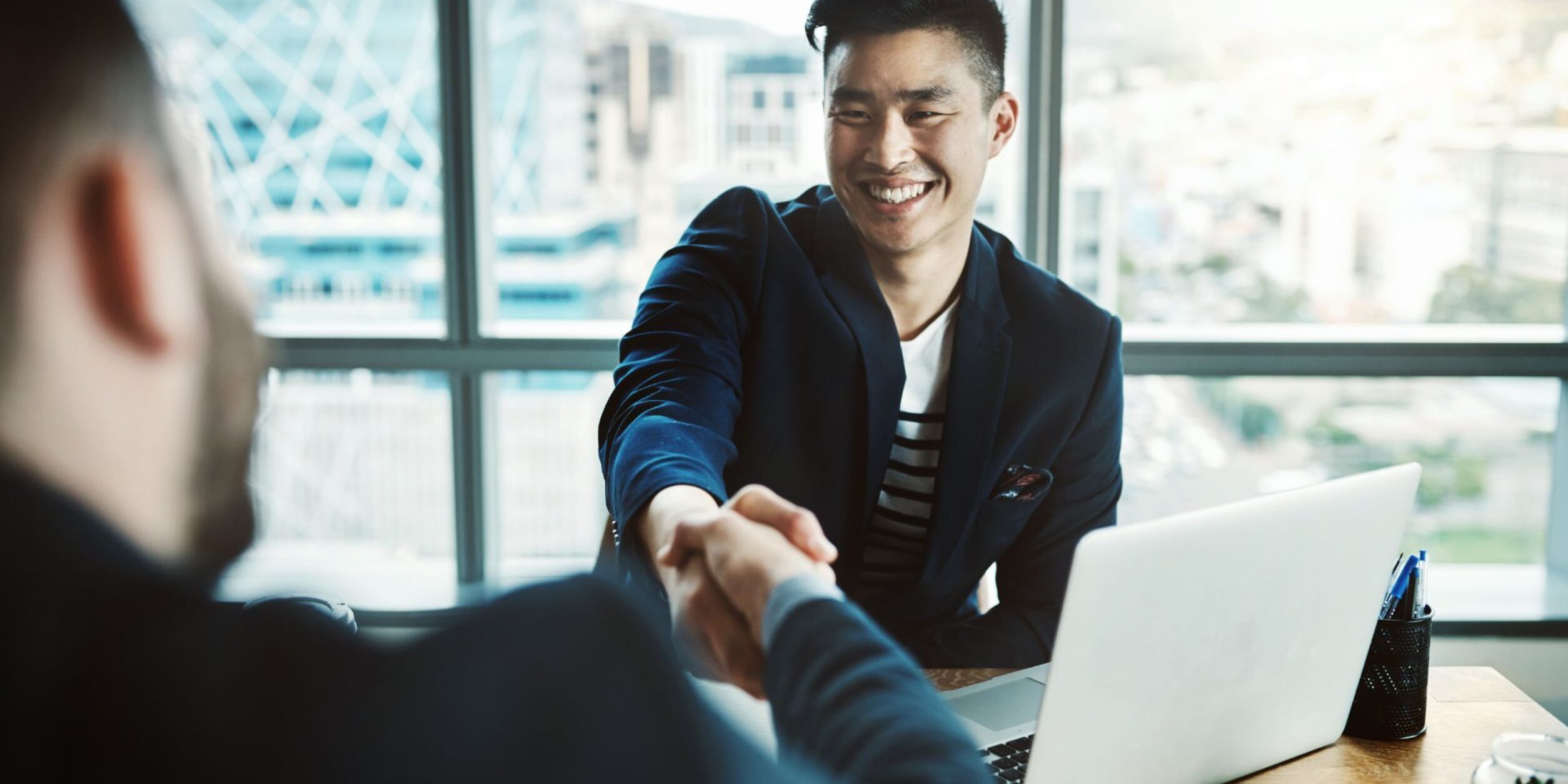 2 Personen sitzen sich am Schreibtisch gegenüber und schütteln sich die Hand
