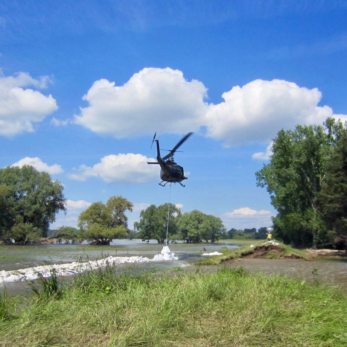 Ein Hubschrauber seilt einen Sandsack an einer Hochwasserbarriere ab.