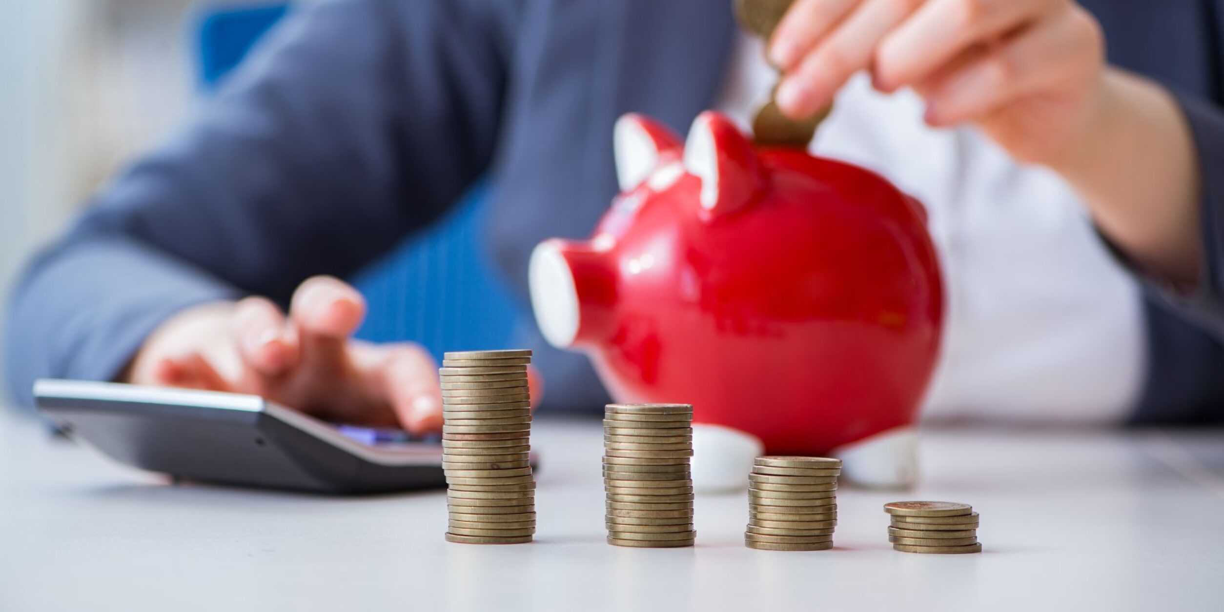 Eine Person wirft Geld in ein Sparschwein, davor sind Münzstapel aufgereiht