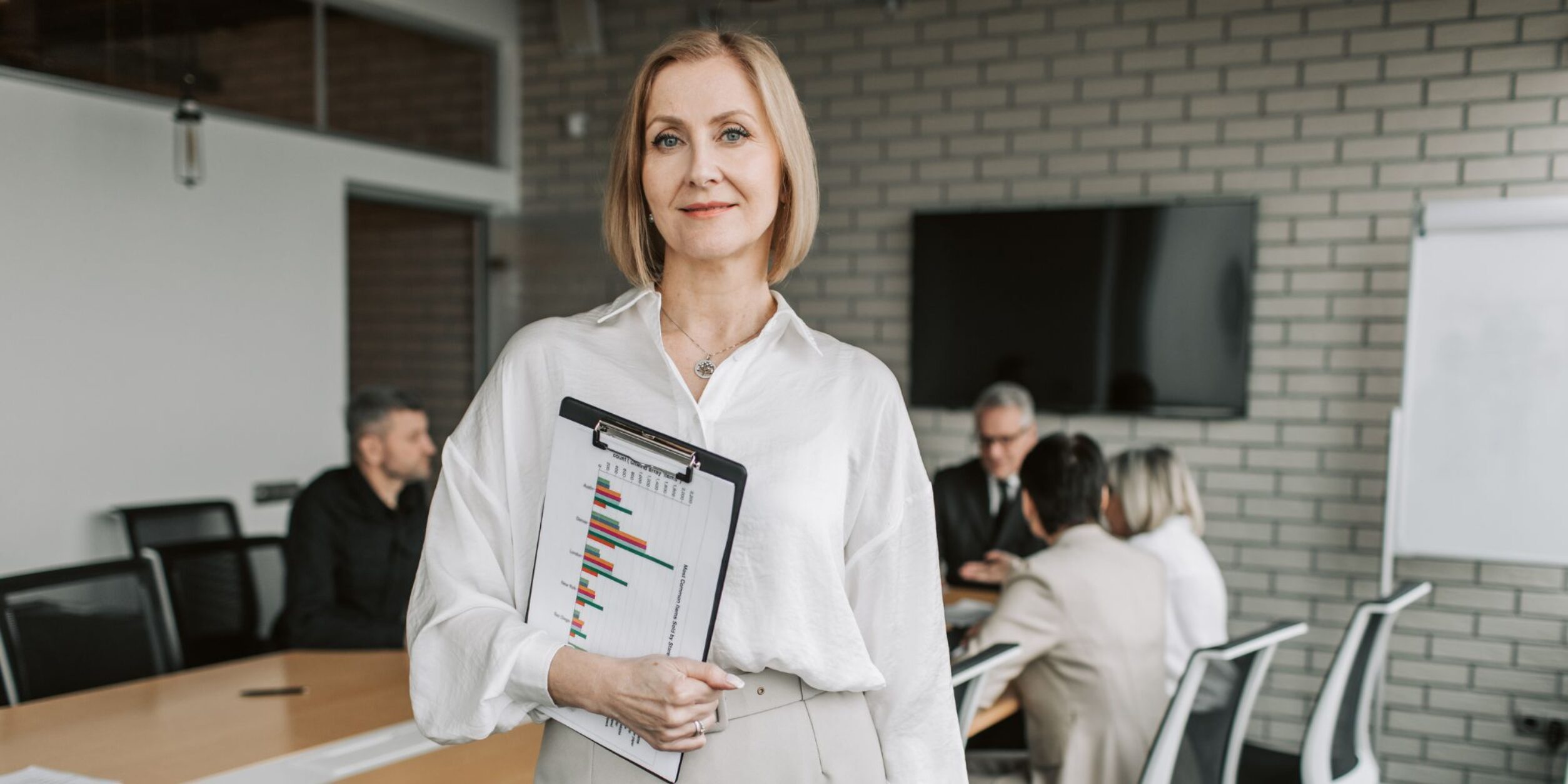 Eine Frau steht mit Klemmbrett vor einem Meetingtisch und blickt in die Kamera