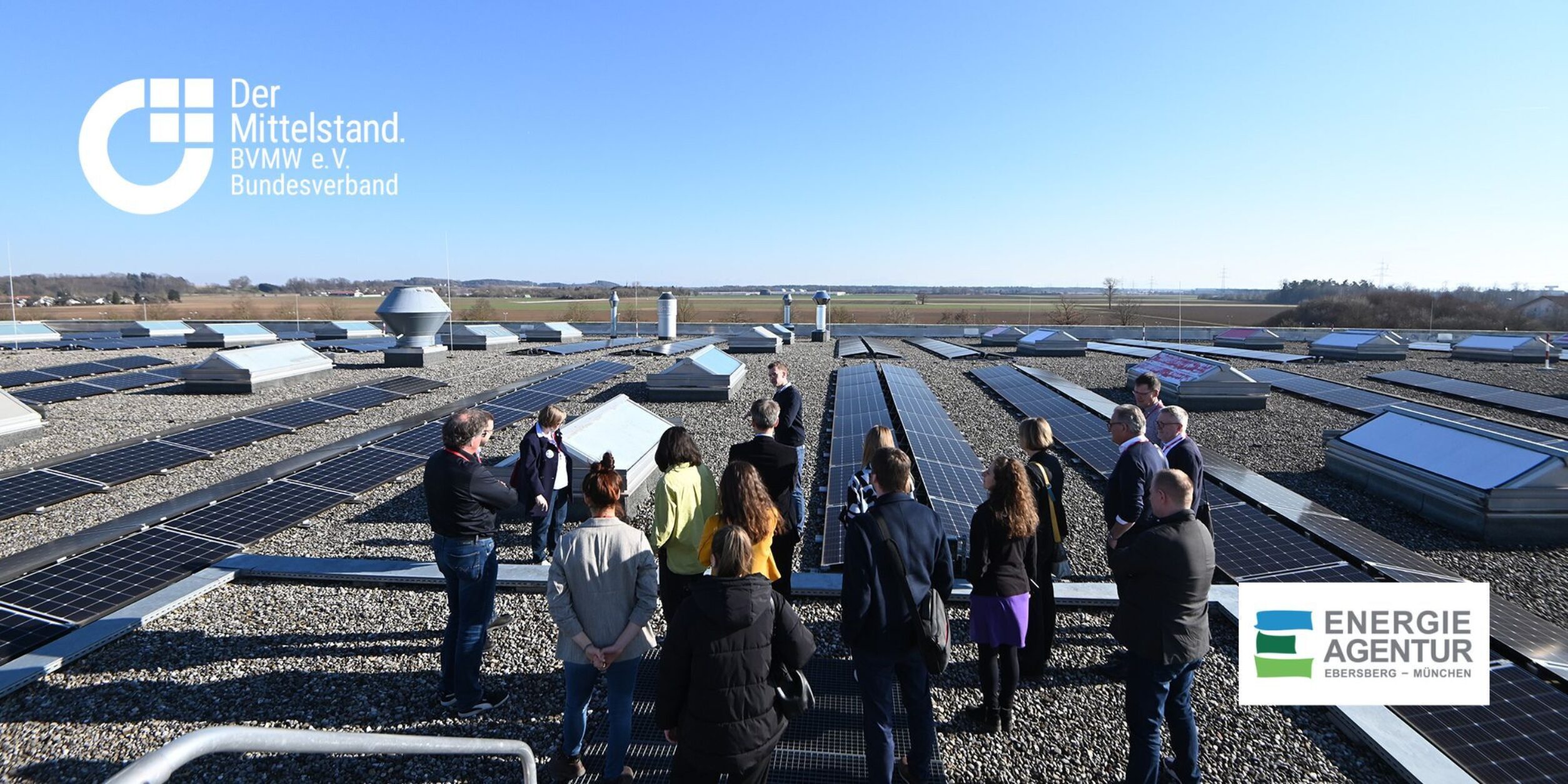 Menschen stehen auf Flachdach mit Solarkollektoren, Logo BVMW e.V., Logo Energieagentur Ebersberg