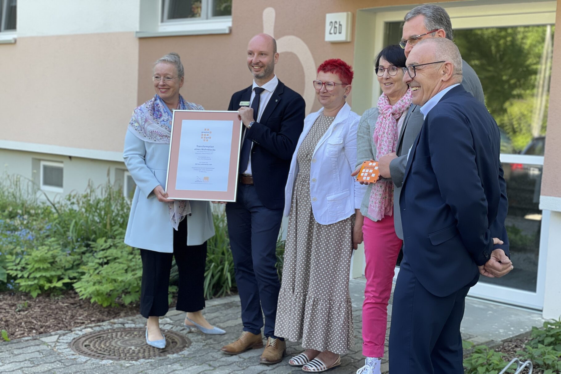 Meschan stehen vor einem Wohnblock und halten eine Urkunde in die Höhe