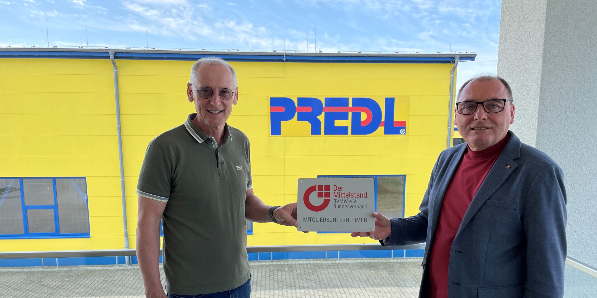 zwei Männer stehen vor einem Firmengebäude und halten ein Schild in die Höhe