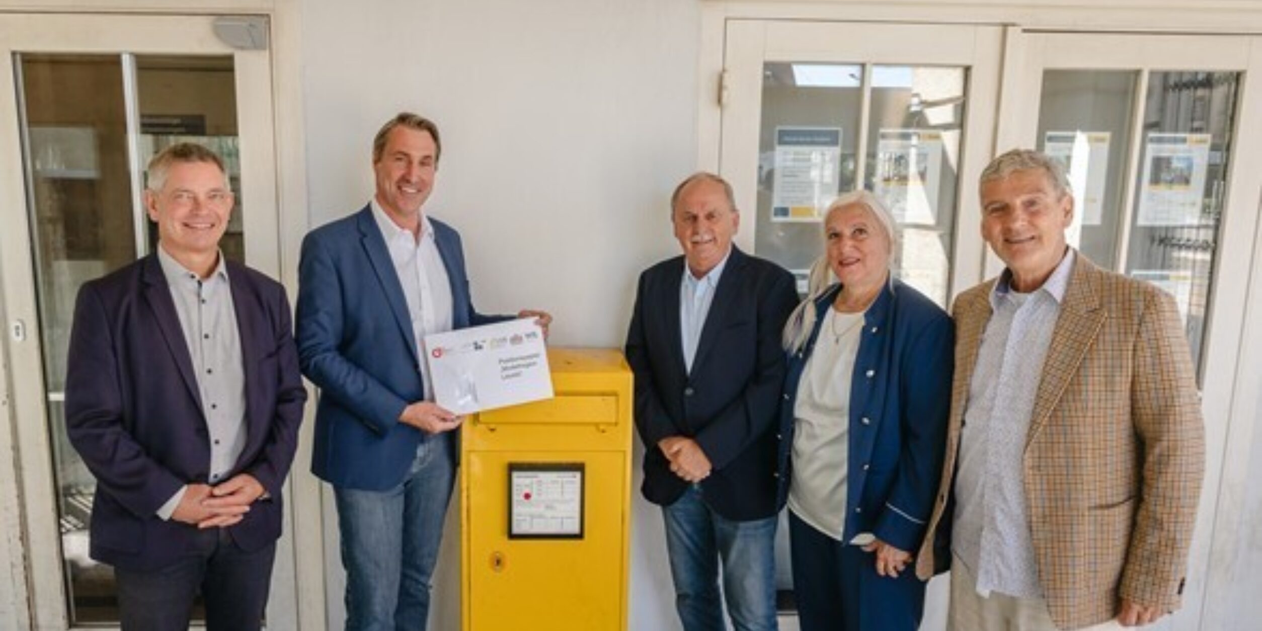 Vier Männer und eine Frau stehen mit einem Brief vor einem Briefkasten