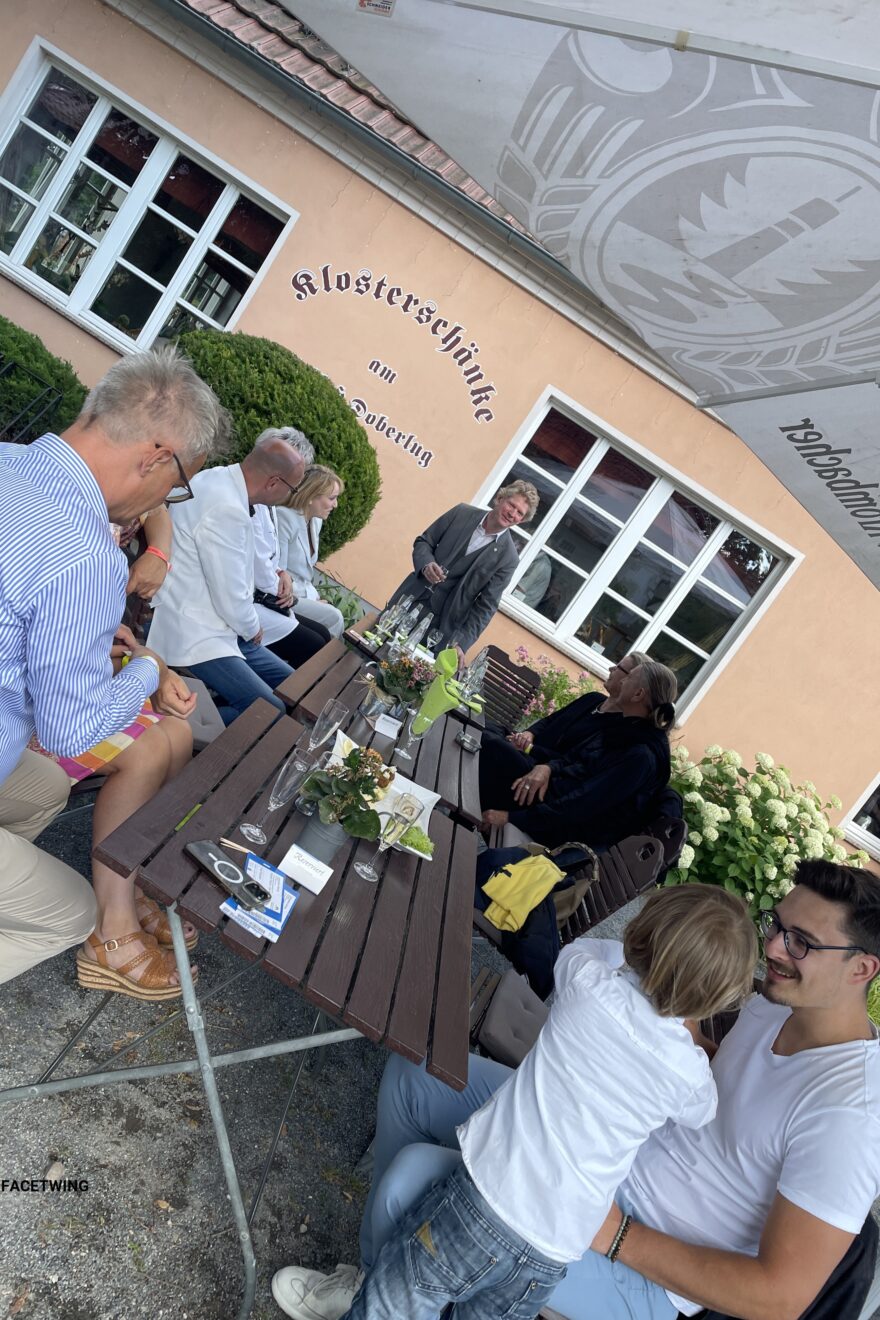 Unternehmer*innen sitzen im Biergarten und werden durch einen Mann begrüßt