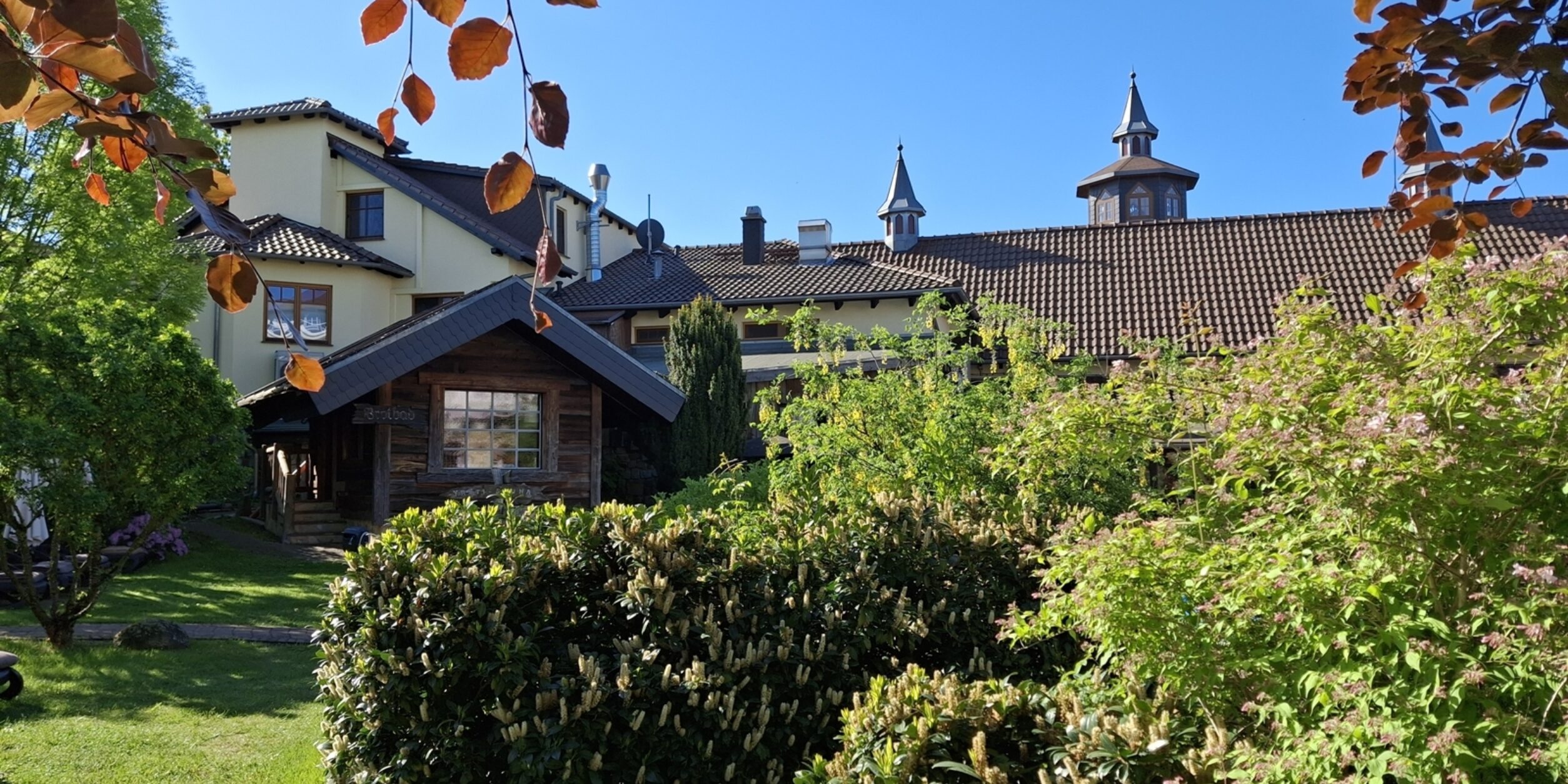 Blick durch den Spa-Garten zum Wellnesshotel Seeschlösschen. Grüne Büsche stehen im Vordergrund. Rechte Seite ein grüner Baum. Im Hintergrund ist das Gebäude des Hotels zu sehen mit kleinen Türmchen und mit verwinkelter Baustruktur. Oben blauer Himmel und herabhängende orangerote Blätter an Ästen eines Baumes.
