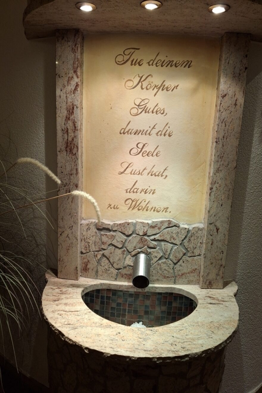 Ein Brunnen an der Wand mit Schrifttafel, welche den Text hat: " Tue deinem Körper Gutes, damit die Seele Lust hat, darin zu Wohnen." Ein Wasserrohr kommt aus der Wand und führt in ein Becken.