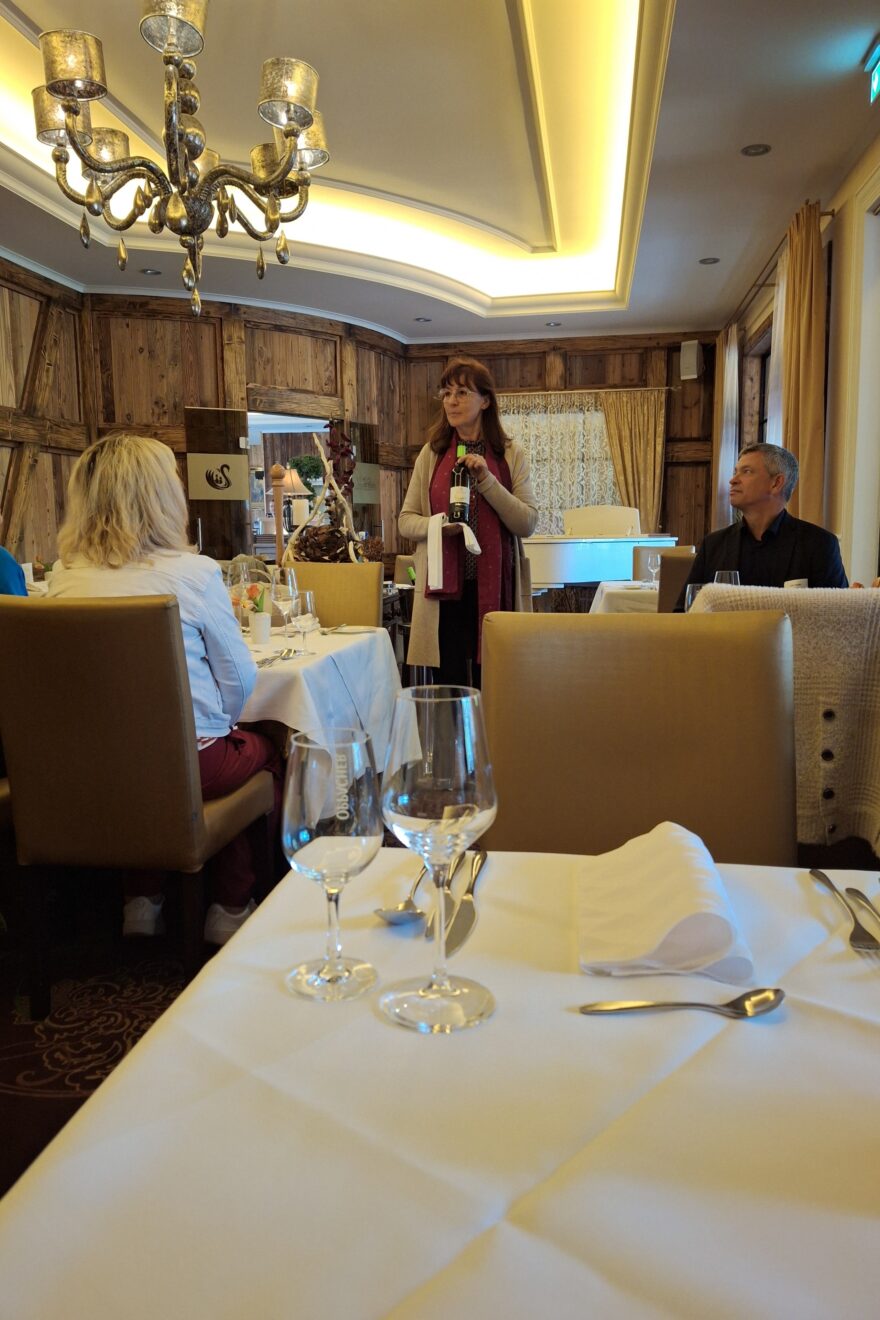 Mit Holzvertäfelter Raum mit Tischen und Stühlen. Teilnehmer der Veranstaltung "Der Tag der guten Seelen" sitzen und sehen zu einer Frau die in der Mitte des Raumes mit einer Weinflasche steht. Auf den Tischen stehen Weingläser, Servietten, Besteck und Geschirr für ein zwei Gänge Mittagessen.