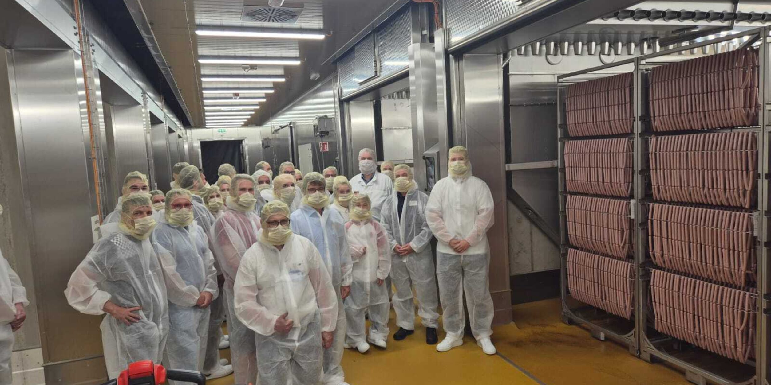 links im Bild: Teilnehmer der Veranstaltung in Hygienebekleidung, rechts im Bild: 1 von 8 Räucherkammern mit 350 000 Würsten