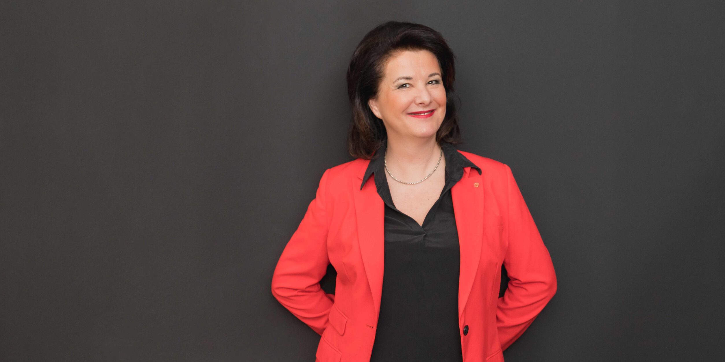 Edda Schmidt in einem roten Blazer vor einer grauen Wand