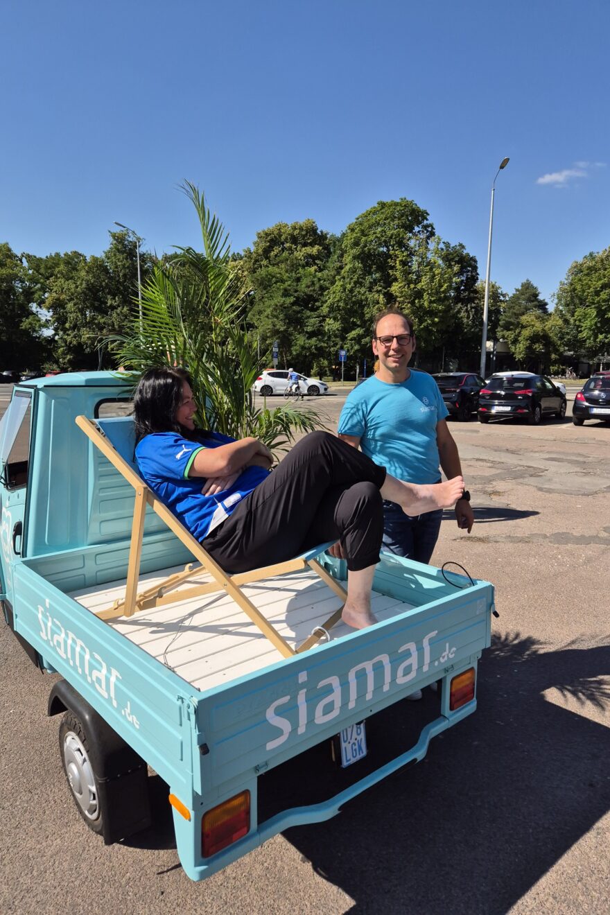 Ein Türkieses Ape (Auto mit drei Rädern) auf der Ladefläche ein Sonnenstuhl mit einer Frau und einem Mann daneben. Aufschrift auf dem Auto ist Siamar