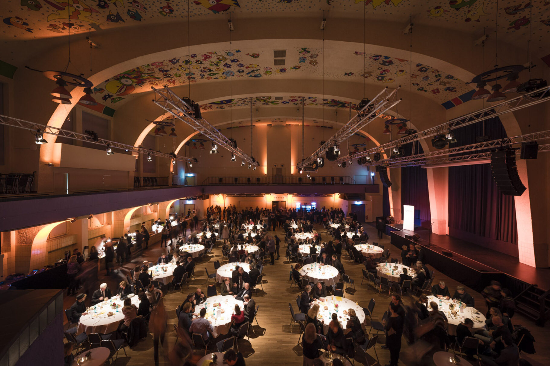 großer Saal mit runden eingedeckten Tischen mit Stühlen und vielen Menschen, Hohe Decke und im Hintergrund eine lange Bar