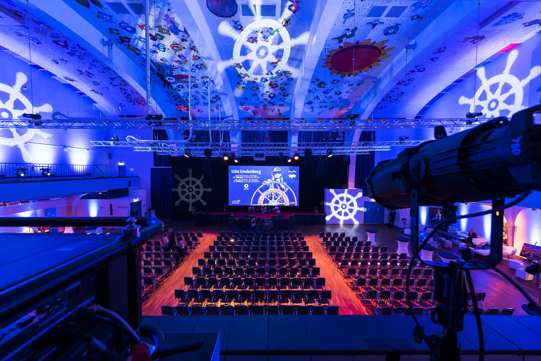 Großer Saal mit vielen Stühlen und roten Teppich mit blauem Licht und Schiffslenkrädern als Lichtern an Decken und Wänden, in der Mitte eine Bühne mit Hintergrund Udo Lindenberg