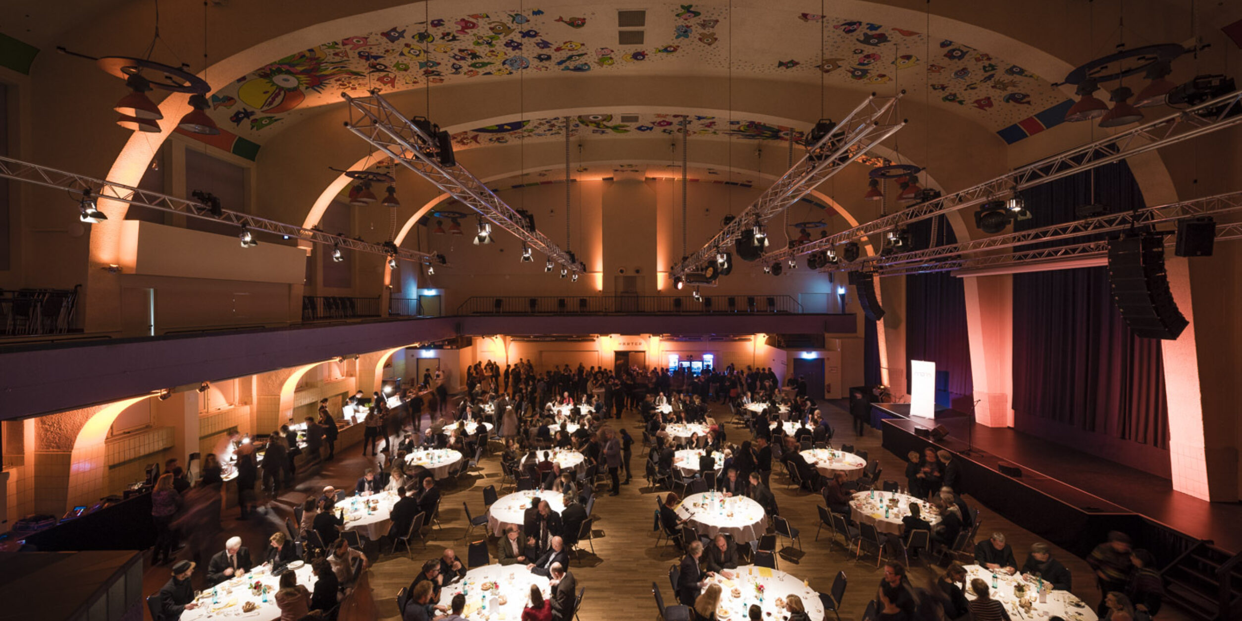 großer Saal mit runden eingedeckten Tischen mit Stühlen und vielen Menschen, Hohe Decke und im Hintergrund eine lange Bar