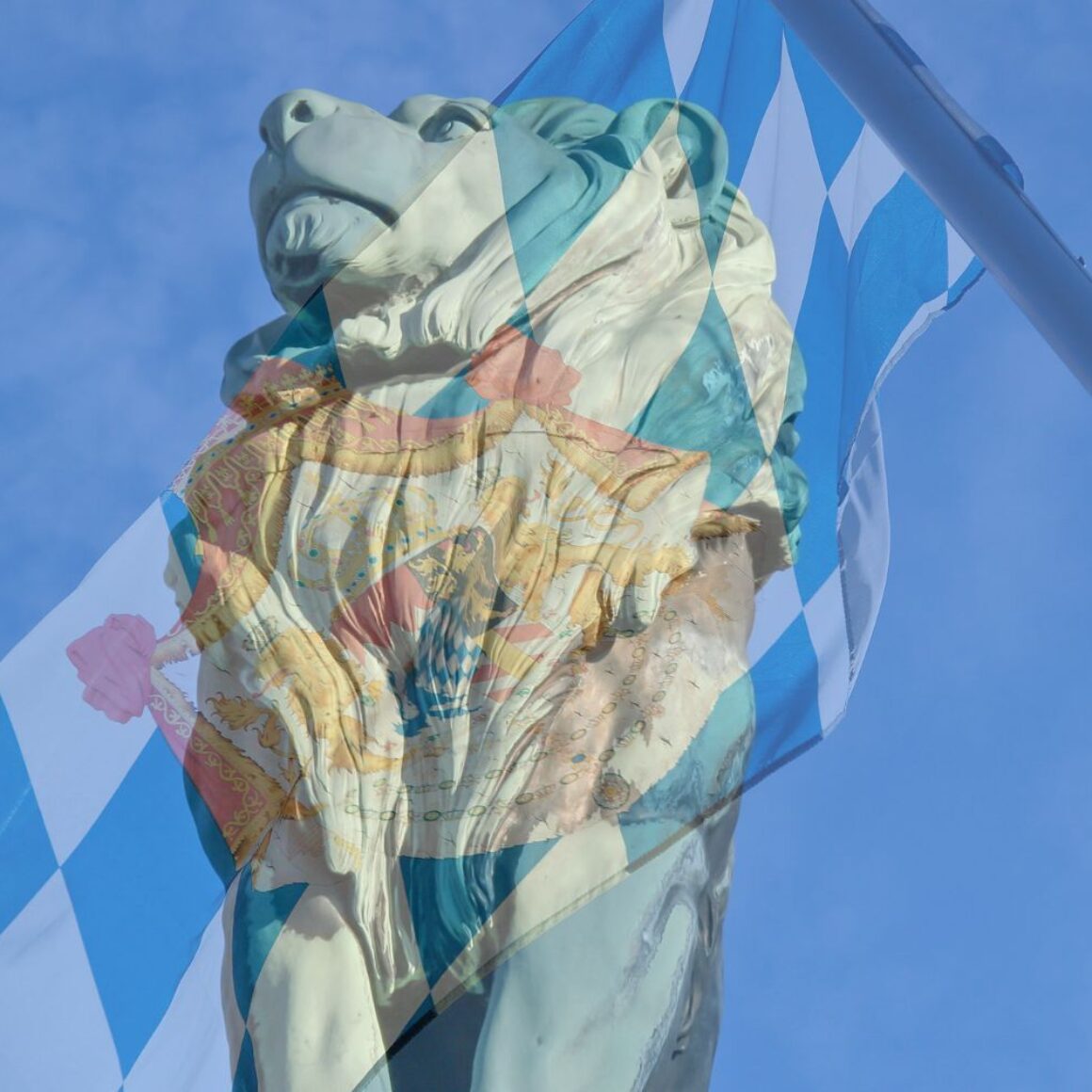 Bayerischer Löwe (Statue) vor blauem Himmel, bayerische Flagge im Hintergrund