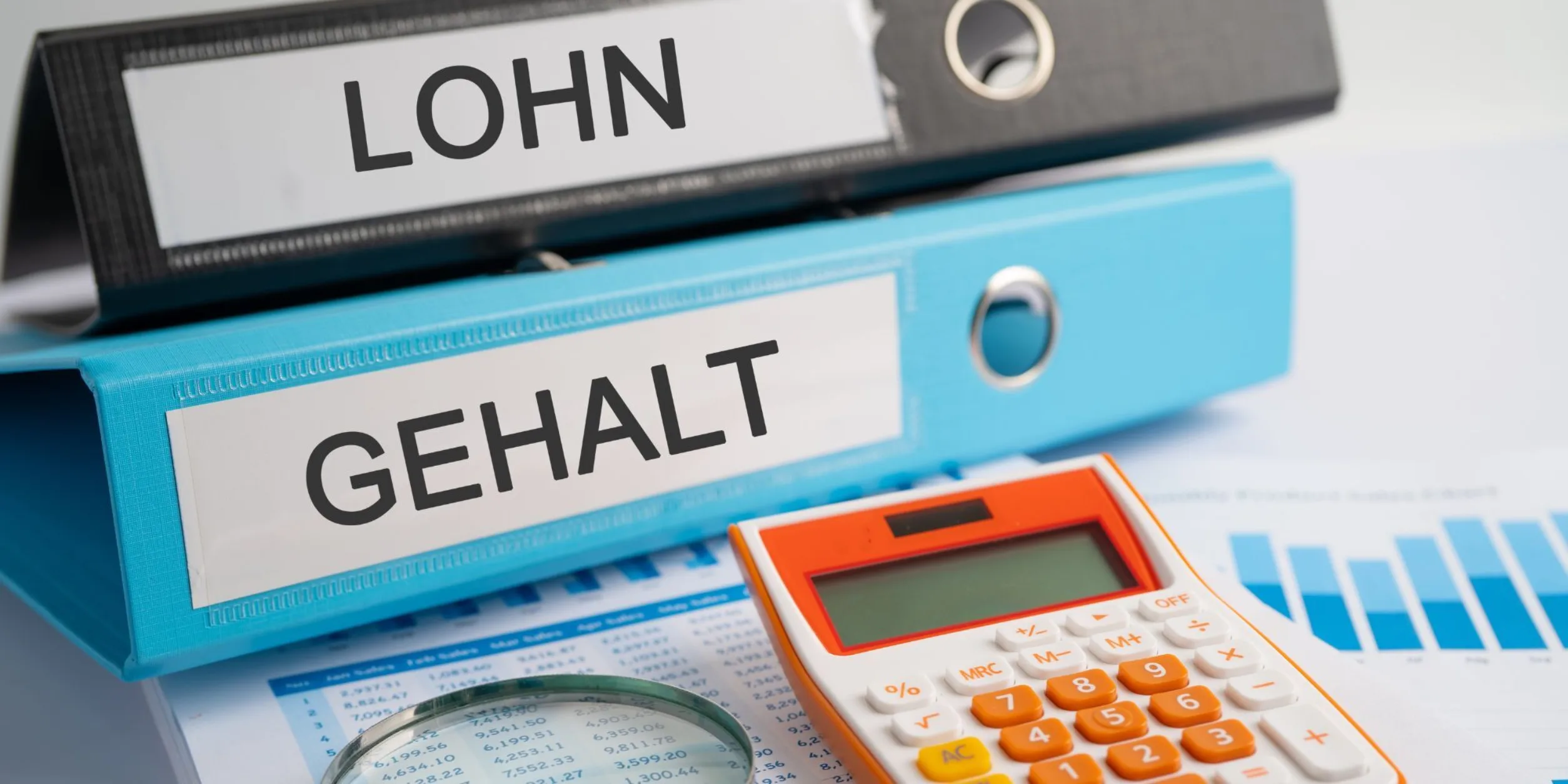 Schwarzer Ordner m. Aufschrift LOHN und blauer Ordner mit Aufschrift GEHALT liegen auf Tisch mit Lohnabrechnung, Lupe und Taschenrechner