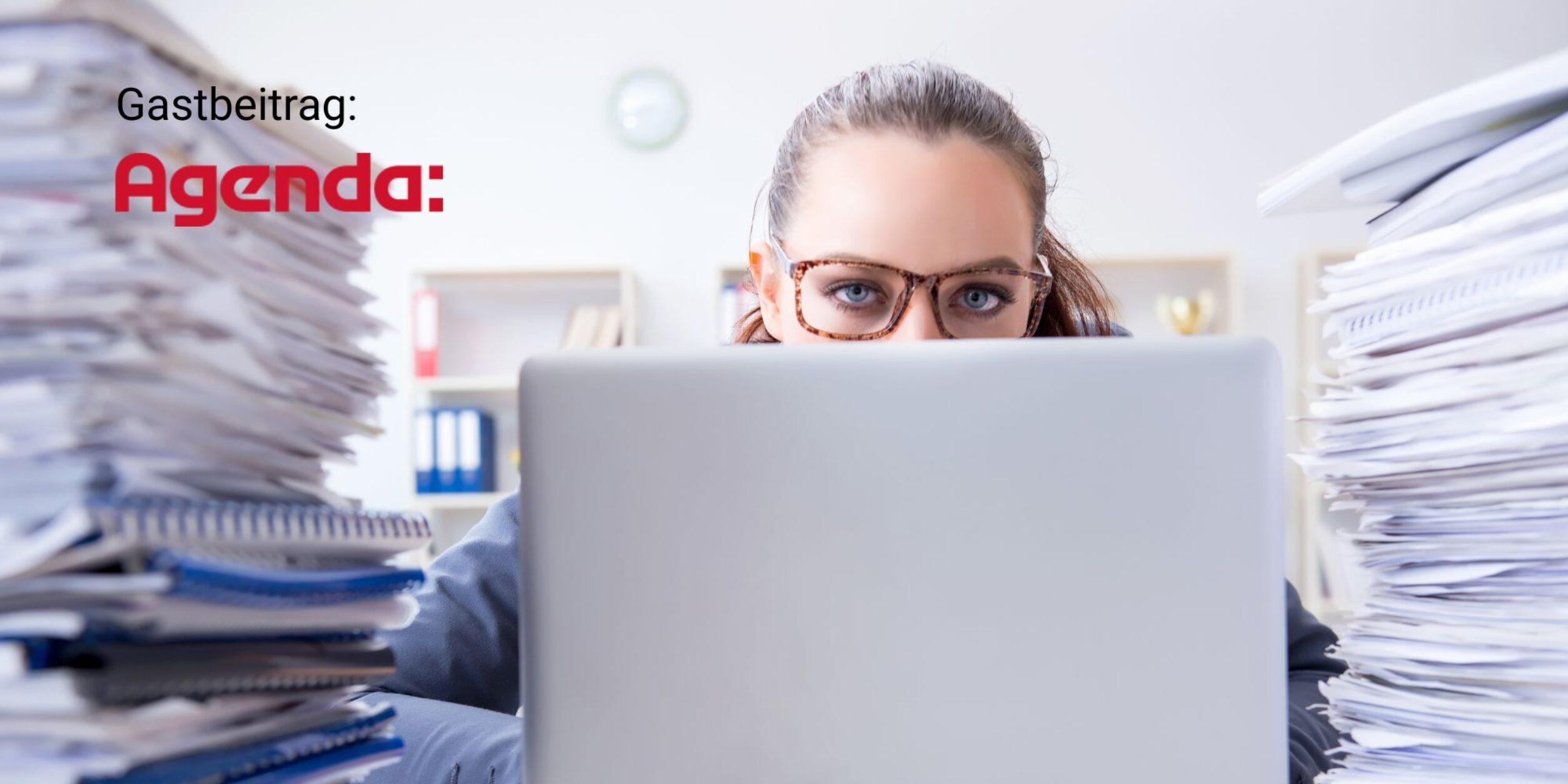 Frau hinter Laptop mit zwei Dokumentenstapeln links und rechts schaut über den Rechner-Deckel