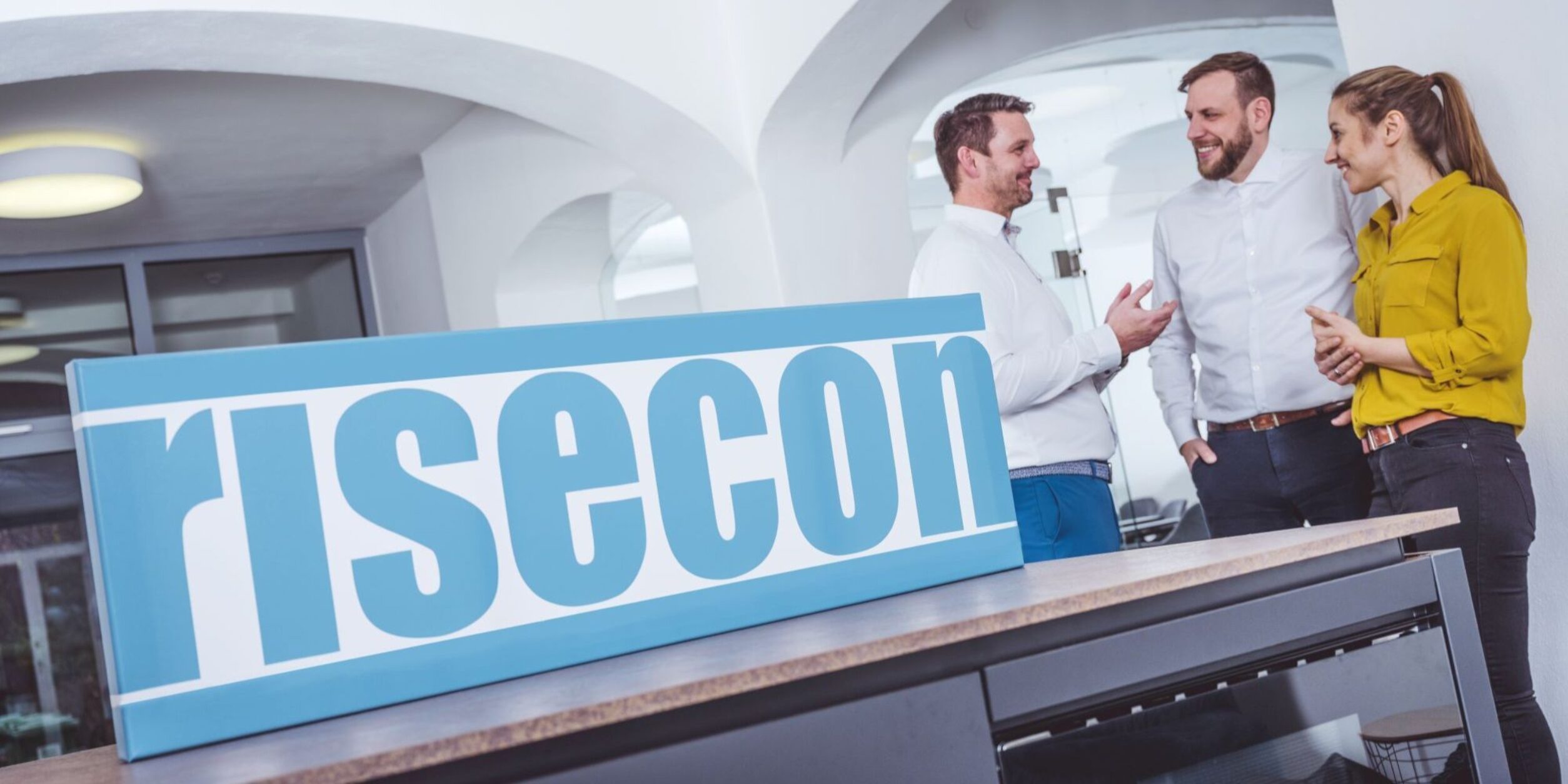 Schriftzug in hellblau "risecon", zwei Männer in weißen Hemden, eine Frau mit Pferdeschwanz und gelber Bluse rechts im Bild