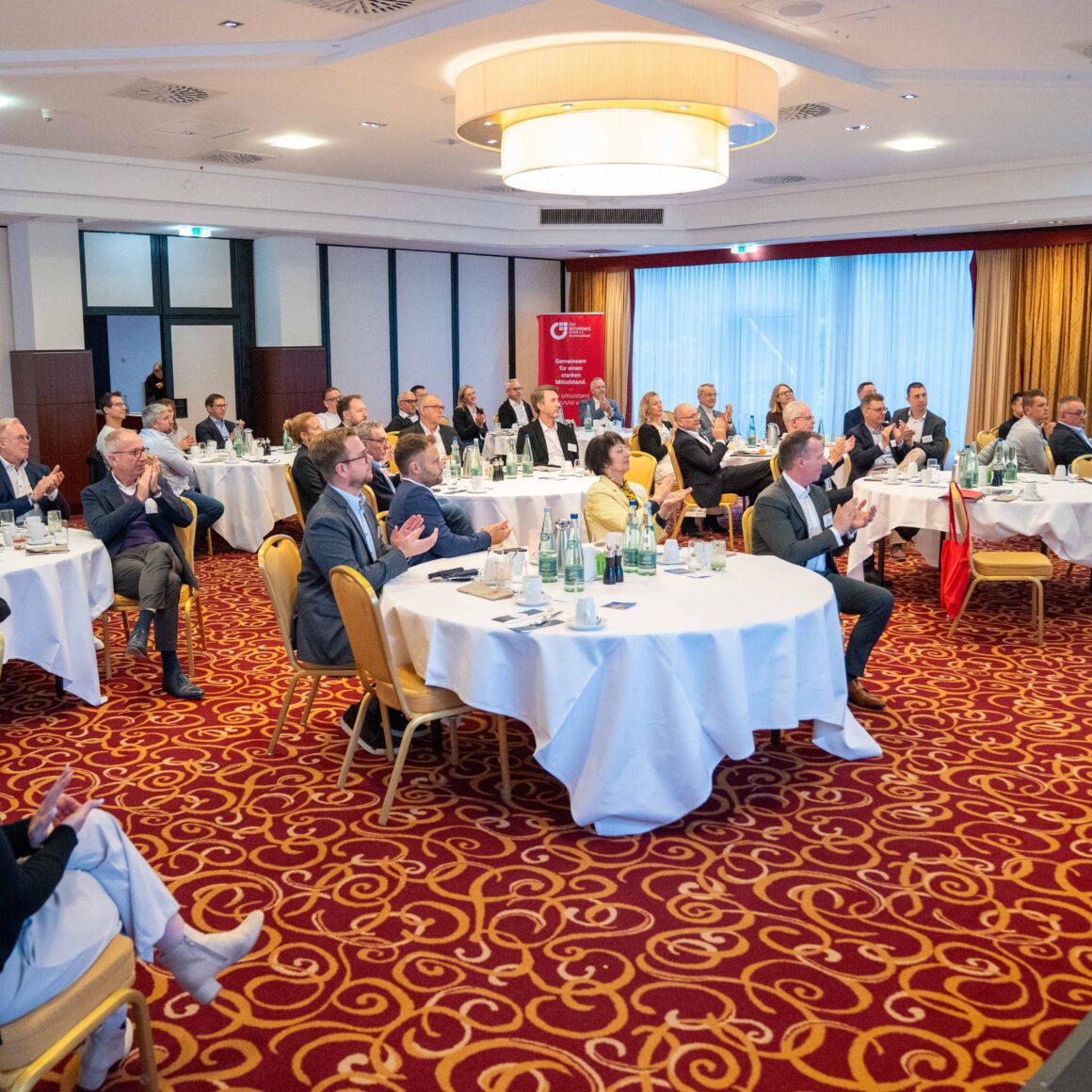 Blick auf Teilnehmer die an Tischen sitzen und klatschen.