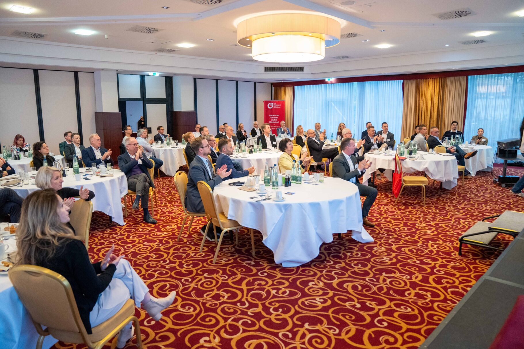 Blick auf Teilnehmer die an Tischen sitzen und klatschen.