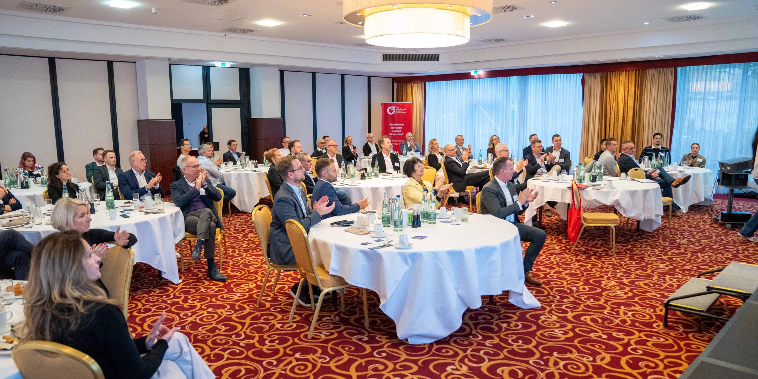 Blick auf Teilnehmer die an Tischen sitzen und klatschen.