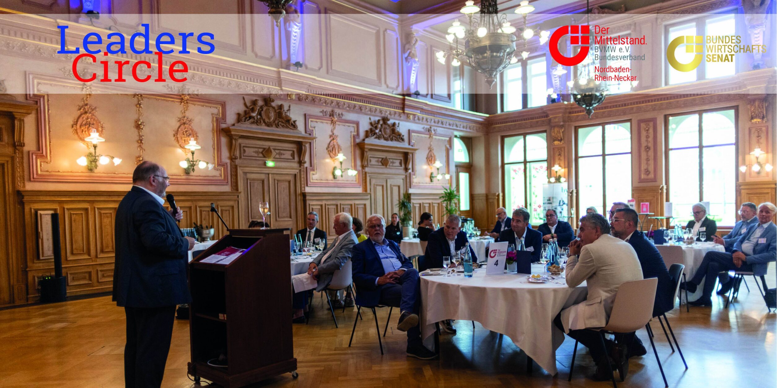 Blick auf die Teilnehmer an ihren Tischen, die der Begrüßung zuhören